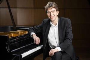 Young Concert Artists - Butte Symphony Young man in a suit smiling while seated at a grand piano in a concert hall setting