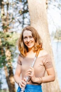 Smiling woman standing outdoors by a lake, holding a silver flute.